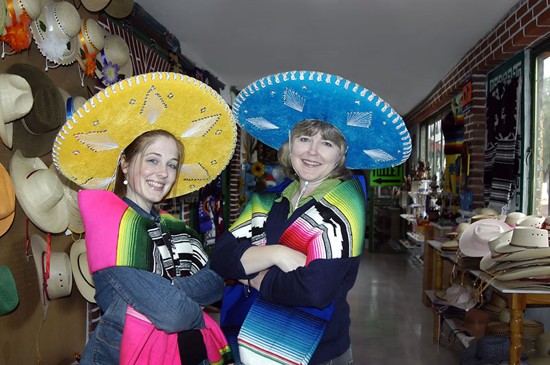 Mother and daughter model Mexican sombreros and colorful blankets. Mother is wearing a bright blue hat and daughter is wearing a yellow. Both tourists are standing in small Mexican Store outside of Mexico City Mexico.